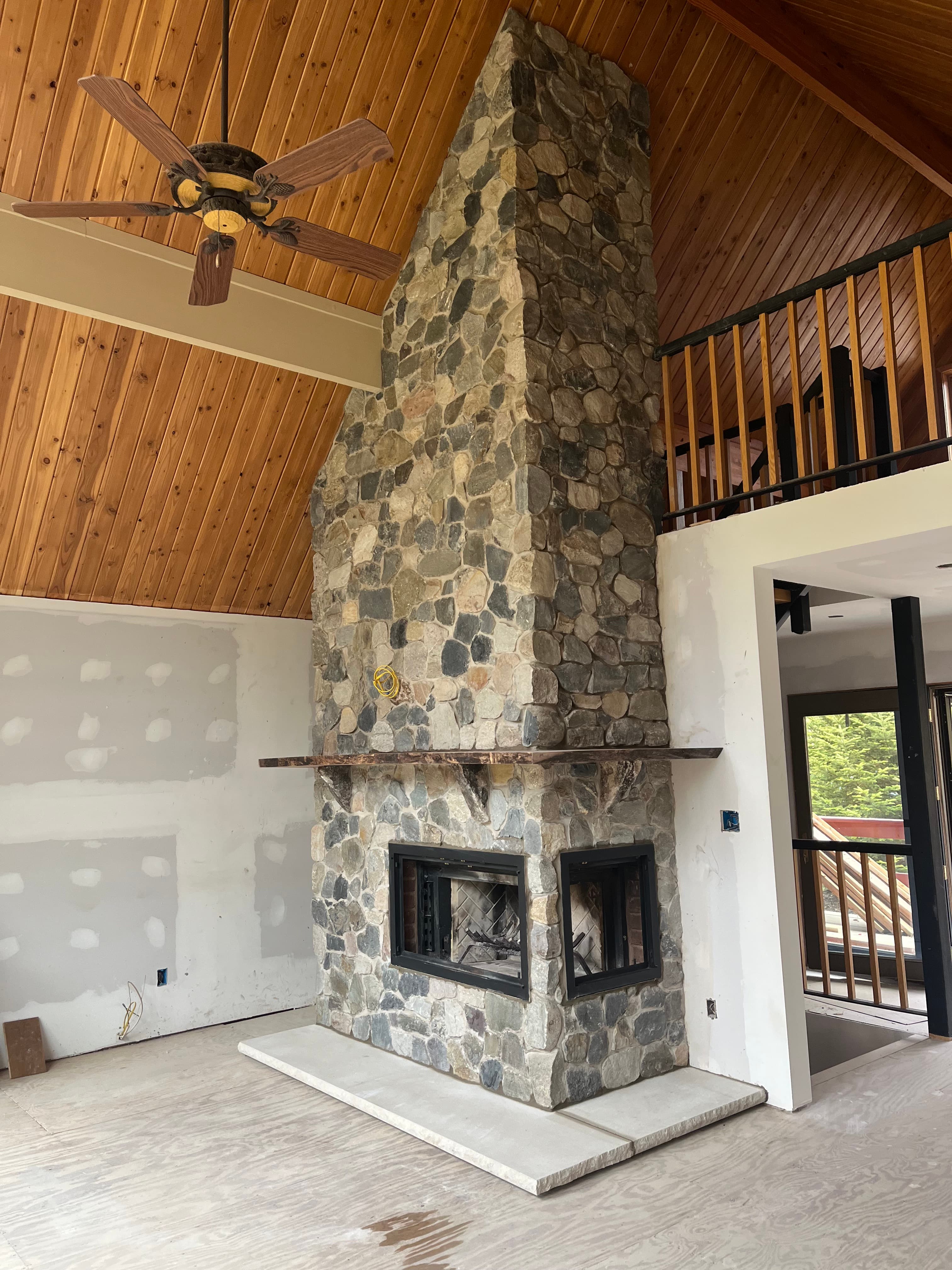Tall stone fireplace in a room with wood-paneled ceilings, a ceiling fan, and stairs.
